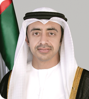 شخص يرتدي الزي التقليدي الخليجي الأبيض مع عباءة سوداء مزينة بخطوط ذهبية يقف أمام علم يحمل ألوان الأحمر والأخضر والأبيض. Description generated by AI
