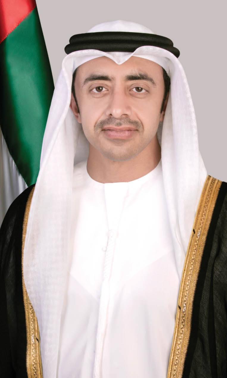 شخص يرتدي زيًا تقليديًا خليجيًا أبيض مع عباءة سوداء مزينة بخطوط ذهبية وخلفه علم دولة الإمارات العربية المتحدة. Description generated by AI