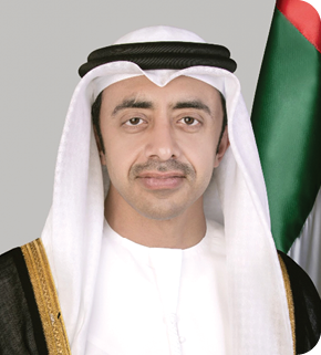 شخص يرتدي الزي التقليدي الخليجي الأبيض مع عباءة سوداء مزينة بخطوط ذهبية يقف أمام علم يحمل ألوان الأحمر والأخضر والأبيض. Description generated by AI
