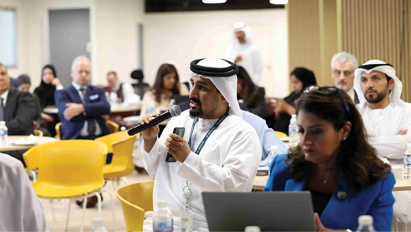 قاعة داخلية واسعة في مبنى حديث تحتوي على أرائك وكراسي مرتبة حول طاولات صغيرة مع وجود أشجار داخلية وأشخاص جالسين ومشيين تحت سقف زجاجي مقوس. Description generated by AI