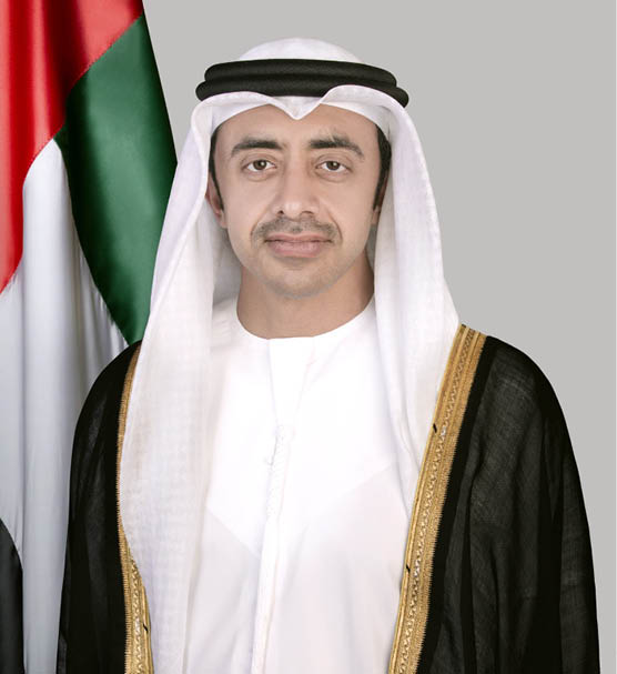 شخص يرتدي زيًا تقليديًا خليجيًا أبيض مع غترة وعقال يقف أمام خلفية رمادية وجزء من علم دولة. Description generated by AI