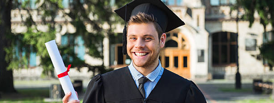 Young male student dressed in black graduation gown. Campus as a background. Boy cheerfully smiling, holding diploma and looking at camera