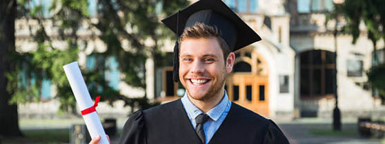 Young male student dressed in black graduation gown. Campus as a background. Boy cheerfully smiling, holding diploma and looking at camera