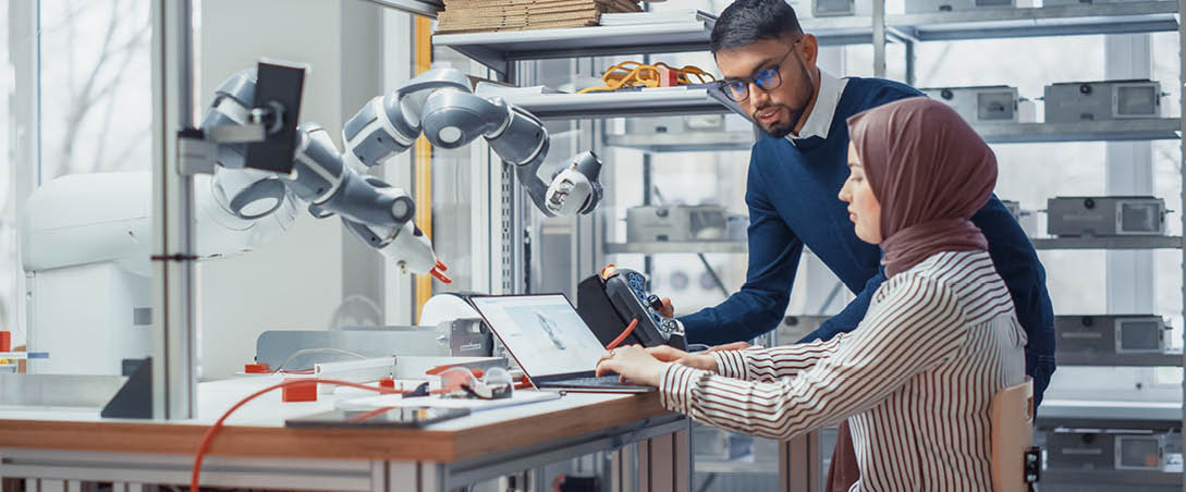 Professional Young Developer in Hijab Coding on Laptop While Her Black Colleague Manipulates Robotic Arm During Factory Workshop. Innovative Technologies And Robotics Concept.