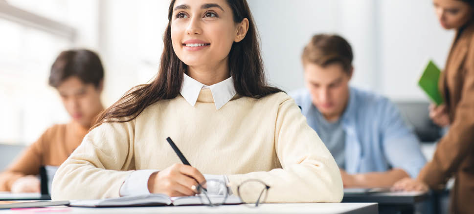 Portrait of smiling female university student holding pen writing in notebook doing examination or quiz test from teacher sitting at desk, attending classroom lesson, looking aside and thinking